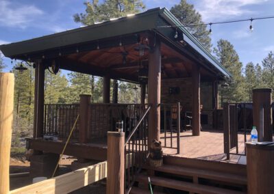 A covered wooden patio with railing stands elevated, featuring a seating area underneath. String lights are hung around, and trees surround the structure. A broom and plastic bottle are in view.