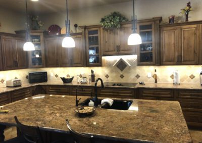 A spacious kitchen with wooden cabinetry, a large island with a granite countertop, overhead pendant lights, and tile backsplash. Various kitchen appliances and decorative items are visible.