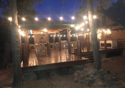 A wooden outdoor deck illuminated by string lights at night, featuring a stone fireplace, seating area, and surrounded by trees.