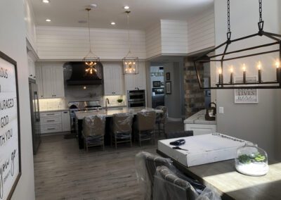 A modern kitchen with white cabinetry, stainless steel appliances, a large island with bar stools, hardwood flooring, and overhead pendant lighting. The dining area features a rectangular table and chandelier.