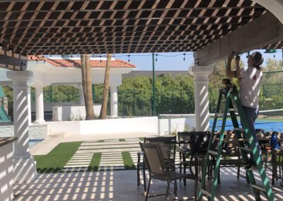 A man stands on a green ladder working on the wooden ceiling of a pergola in a sunlit outdoor area, with a patio and tennis court visible in the background.