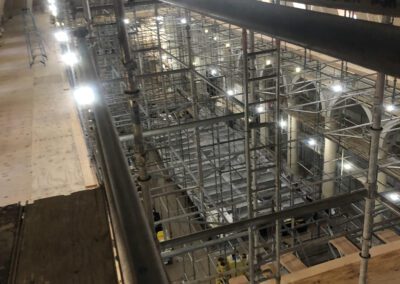 Scaffolding and construction materials set up inside a large hall with arched ceilings and illuminated lights.