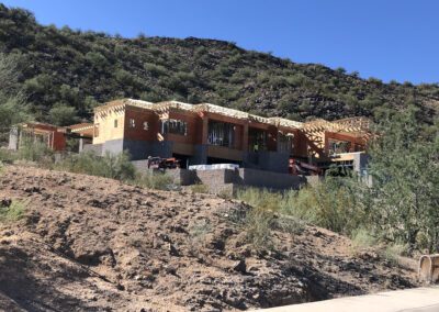 A large house is under construction on a hillside, with wooden frames and walls partially built. Several vehicles and construction materials are visible on the site, surrounded by desert terrain.