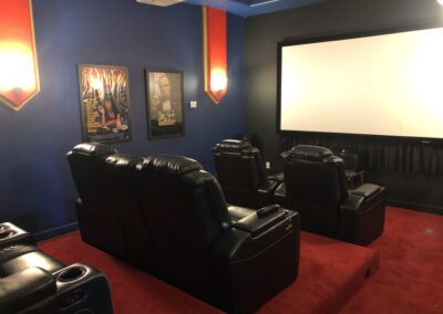 A home theater room with black leather reclining seats, red carpet, a large white screen on a dark wall, and two movie posters on the side wall.