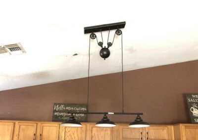 Ceiling-mounted light fixture with three hanging lamps, suspended by cables connected to a pulley system. The background includes a brown wall and a partial view of wooden cabinets.